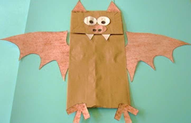 Brown paper bag puppet that resembles a bat. The puppet includes bat wings, eyes, nose, two fang-like teeth, two triangular ears and two feet. The puppet is against a teal blue background. 