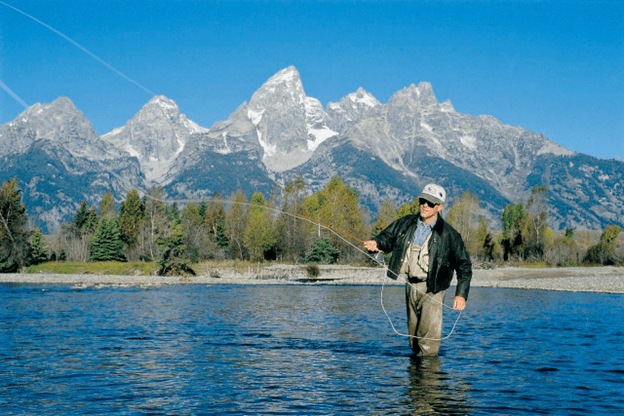 A man is fly fishing in front of the Tetons (stark, striking mountains)